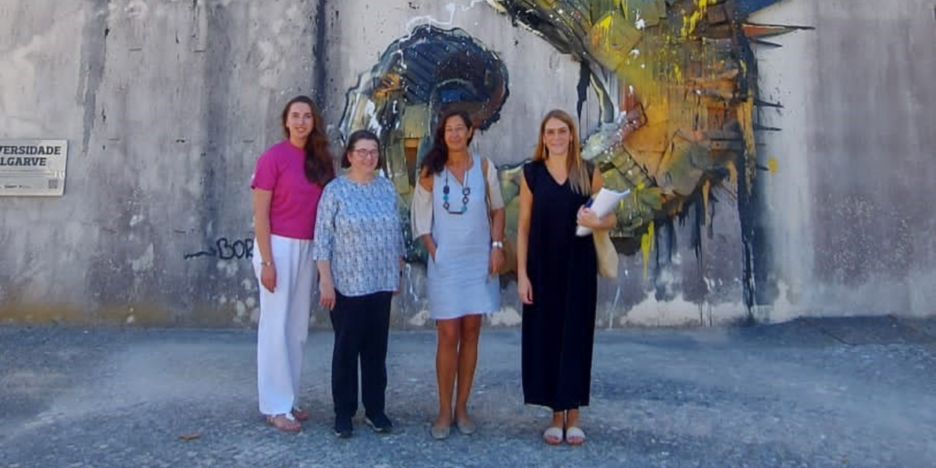 The photo presents four people standing in front of a high façade on which there is an art work presenting a big sea horse which is partly visible in the photo. The colours of the sea horse are yellow and blue, the wall is grey concrete façade. 