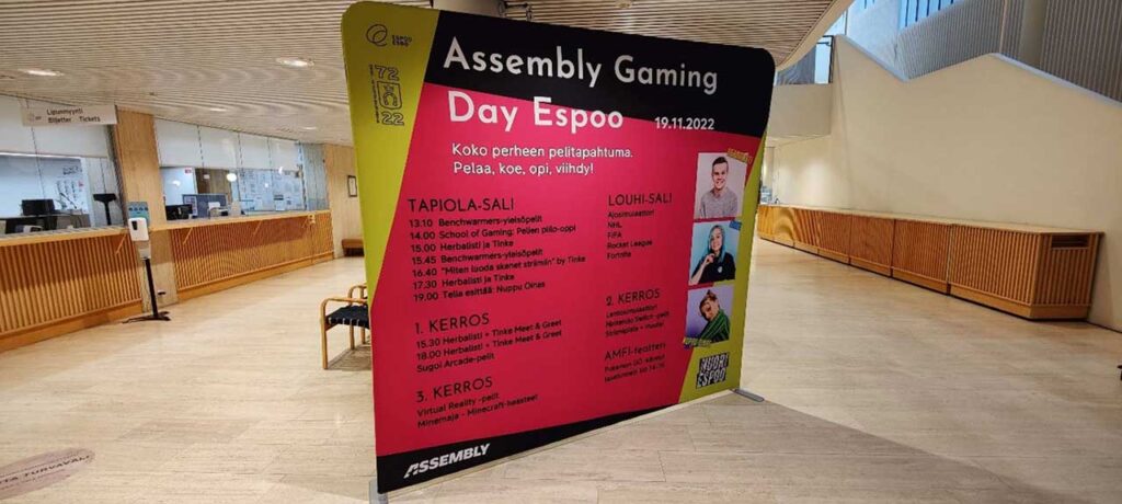 [Alt-teksti: suuren rakennuksen aulassa oleva seisova ohjelmajuliste, jossa teksti Assembly Gaming Day Espoo Koko perheen pelitapahtuma, Pelaa, opi, koe viihdy. Tekstejä myös ohjelmasta tarkemmin Tapiola-salissa, Louhi-salissa ja eri kerroksissa.]