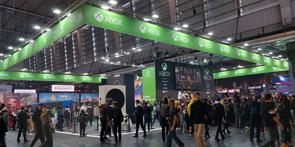 Tapahtumassa Xbox näytteilleasettajan booth, jossa ihmiset jonottavat tai seuraavat Xbox-lähetystä näytöltä. Boothin seiniä koristavat yrityksen logot.]