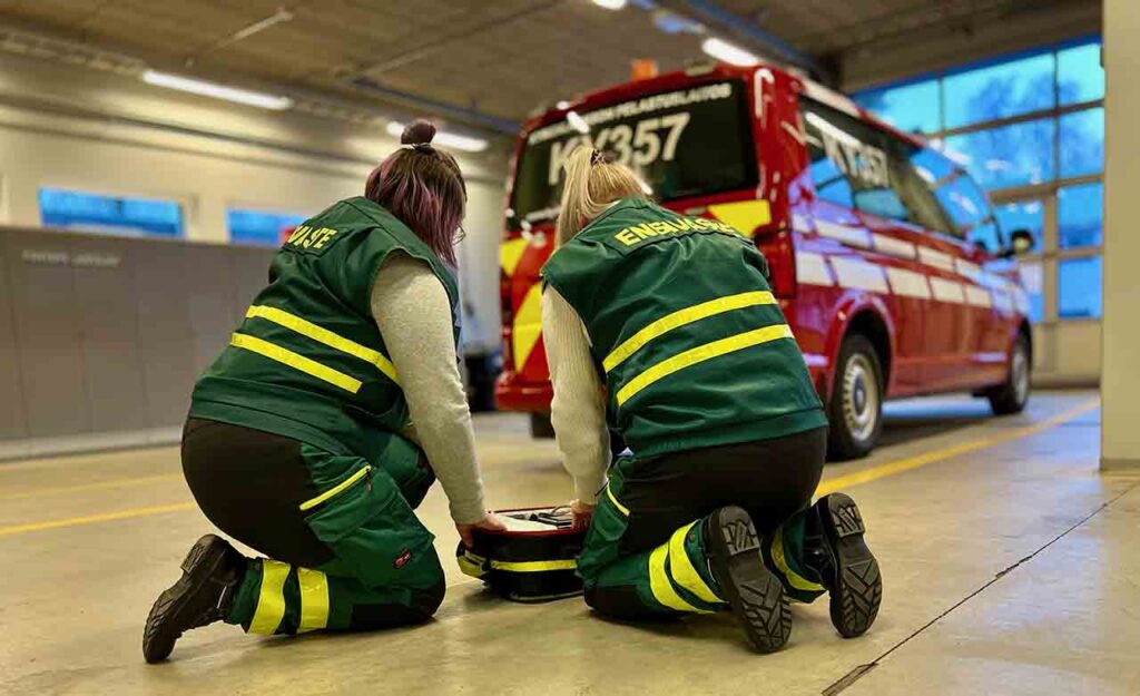 [Alt-teksti: Kaksi naista ensivaste-ammattiasuissa paloaseman tiloissa tutkimassa lattialle asetettua välineistöä.]