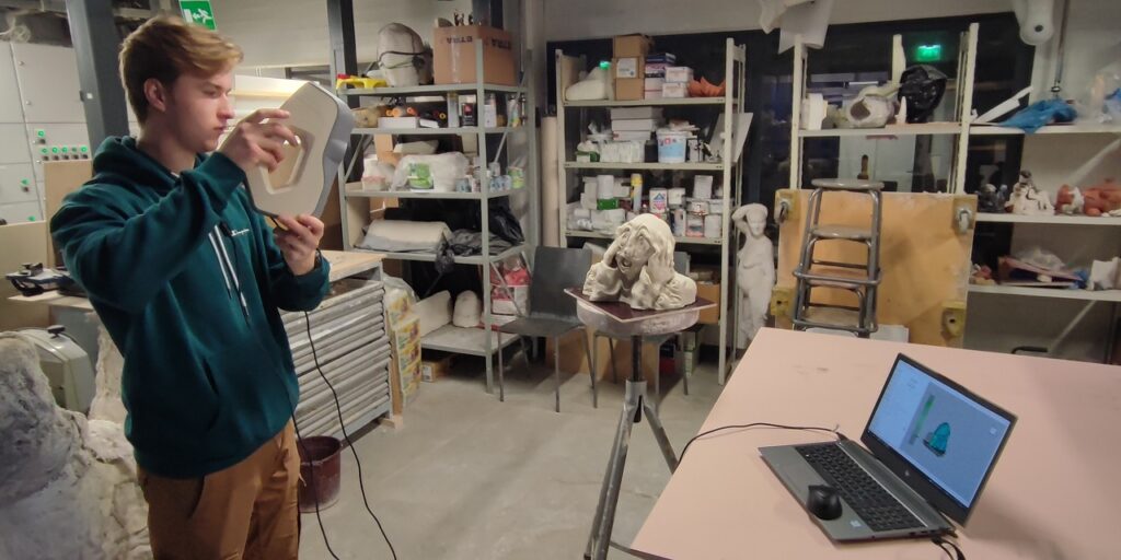 Filled storage room. One person in front in green hoodie, holding scanner in hand, clay sculpture in the back and laptop on the table. 