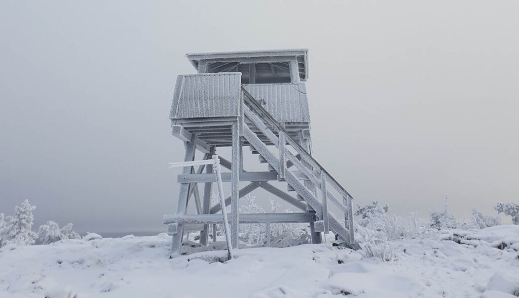 [Alt-teksti: Luminen maisema, mäen laella puinen näkötornirakennelma, johon voi kiivetä portaita pitkin.]