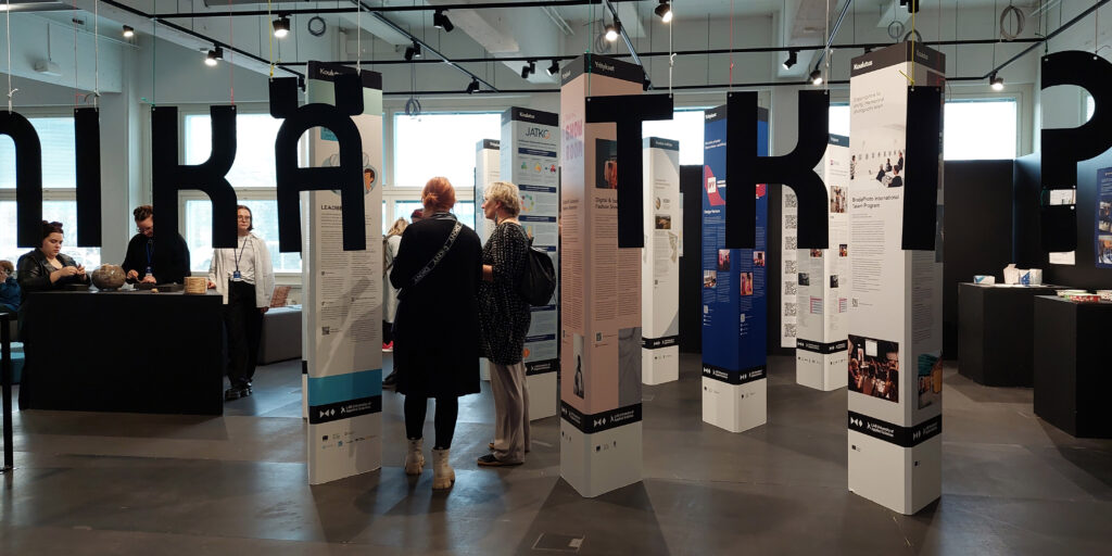 Näyttelytila, jossa näyttelyn nimi: Mikä TKI?, ihmisiä ja näyttelyn hankkeista kertovia infopyloneita. 