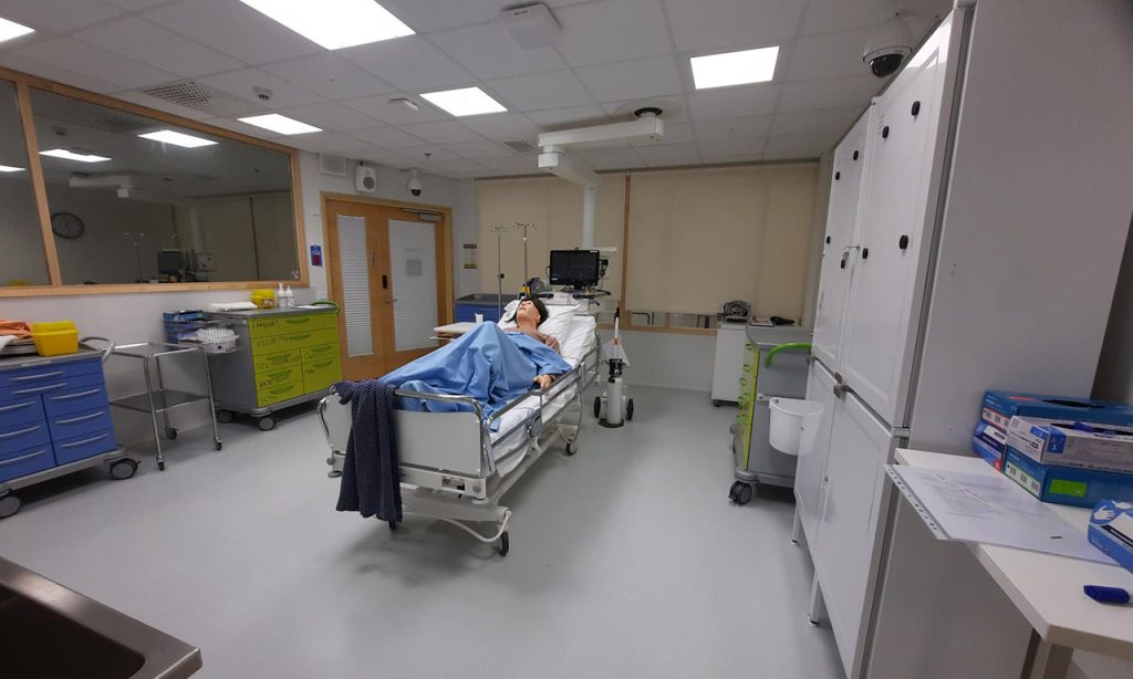 [Alt-teksti: hoitohuone, jonka keskellä on potilassänky. Sängyssä makaa selällään ihmishahmoinen nukke osittain peitolla peiteltynä.]