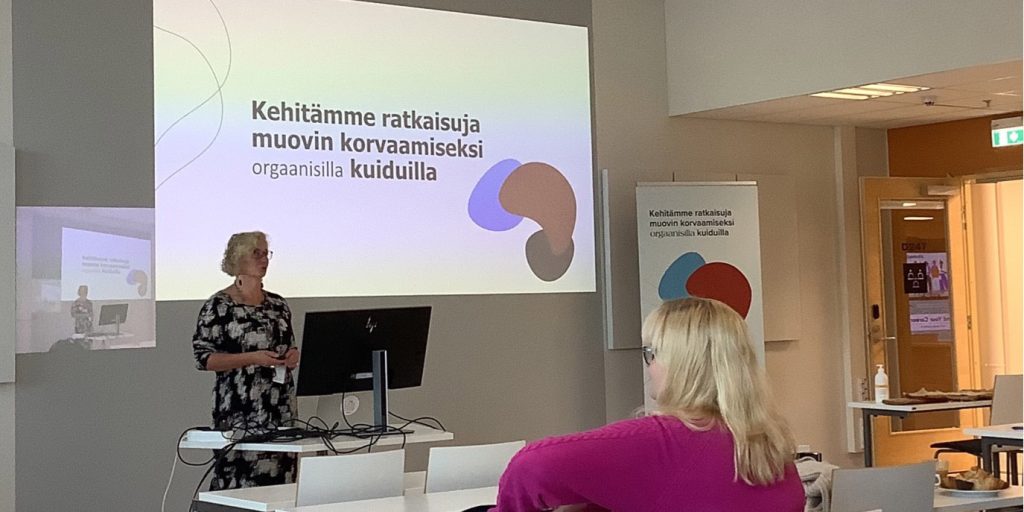 Hiukka 2.0 -hankkeen projektipäällikkö Paula Nurminen seisoo luokan edessä presentoimassa. Kuvan etualalla pöydän ääressä istuu nainen purppuran värisessä paidassa kuuntelemassa presentaatiota. 