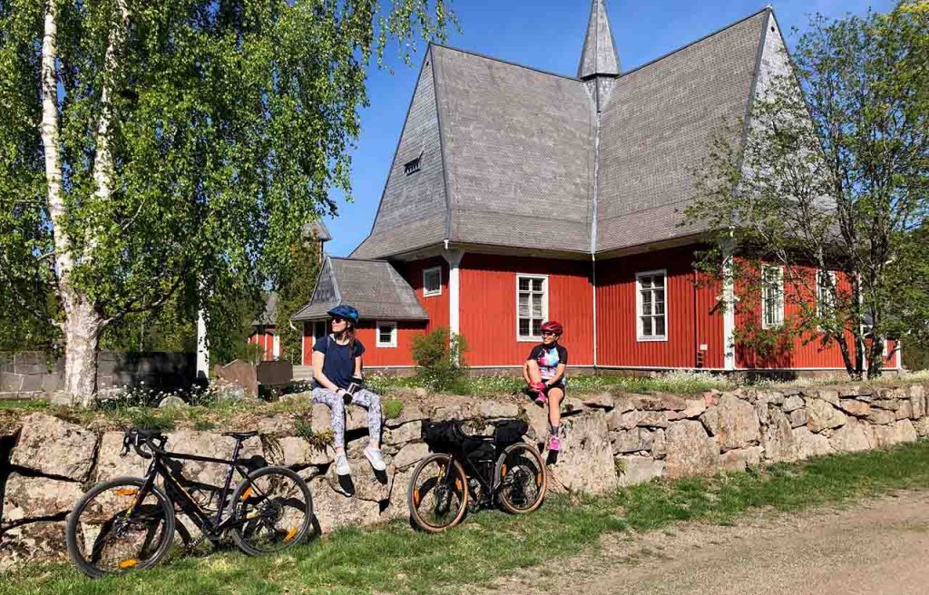 [Alt-teksti: kesäinen päivä ja auringonpaiste, taustalla vanha puinen kirkko ja sen edessä kiviaita, jolla istuskelee kaksi pyöräilijää, heidän pyöränsä nojaavat samaan aitaan.]