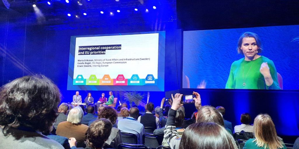 A dark meeting room. People are sitting in rows and watching the stage in front of them. The stage is illuminated with a blue light. There are two big screens, the left screen shows a slide with the title “Interregional cooperation and EU priorities” and some text and colorful icons at the bottom of the slide. The right screen shows a lady in a green dress. On the stage, in the middle, there are four chairs with people sitting on them.
