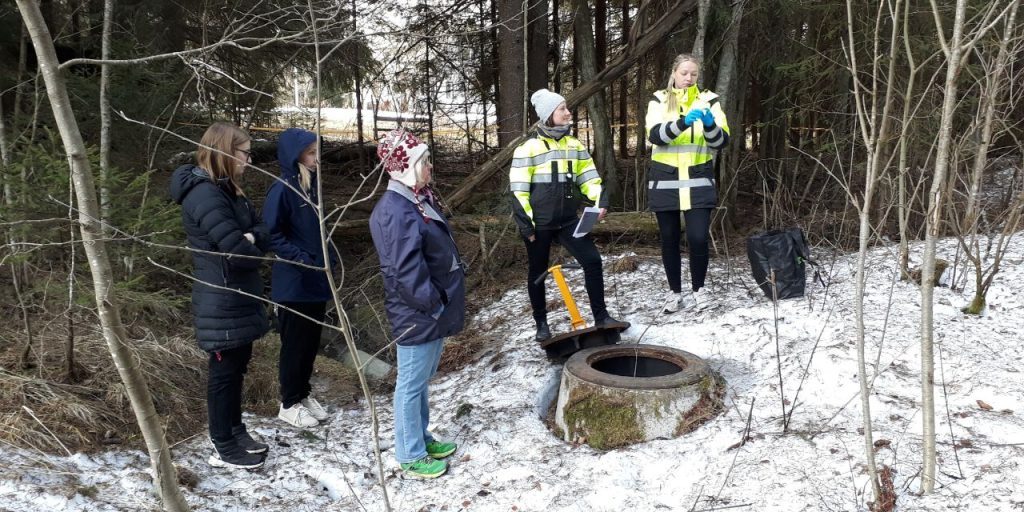Forest on the background, some snow in the ground. Two people in work clothes stand in front of a stormwater well. Three people observe the situation.