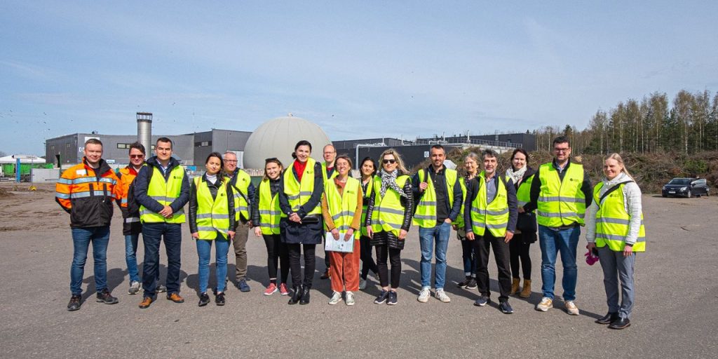 A group of people with yellow safety vests standing outside in an industrial area. At the background, there is a biogas plant.