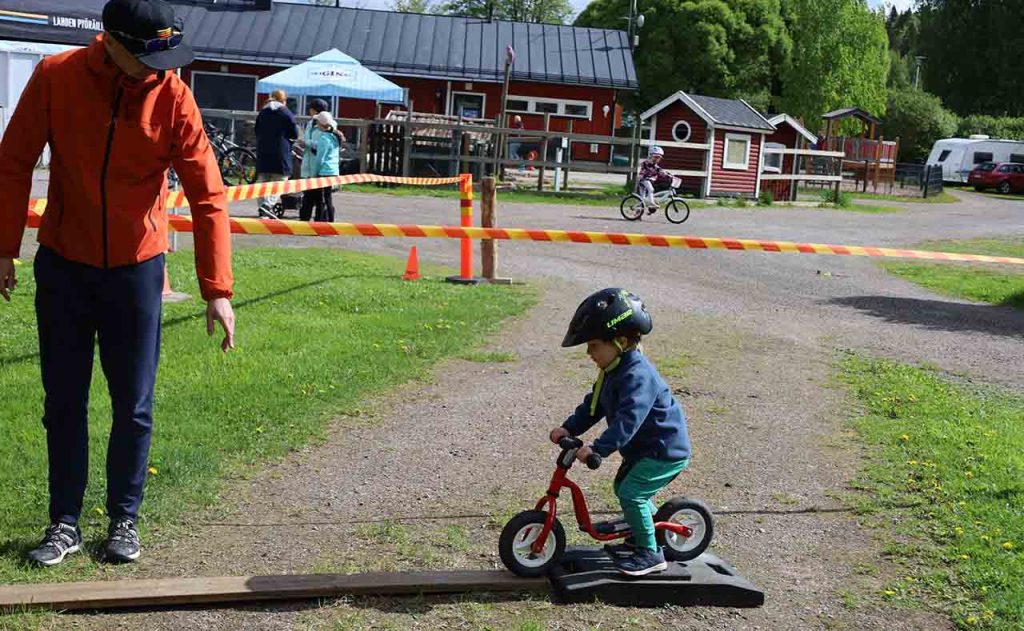 [Alt-teksti: pieni lapsi pienen pyöränsä kanssa, ajamassa maahan asetetulle melko kapealle lankulle, vieressä aikuinen opastamassa.]