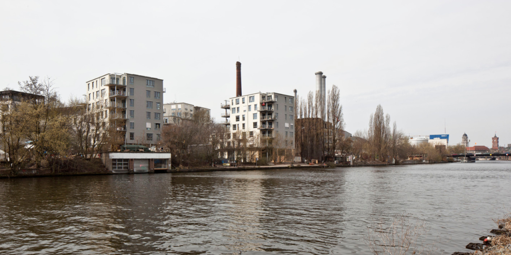 Two apartment buildings seen from the far side of a river and also some chimneys in the background.