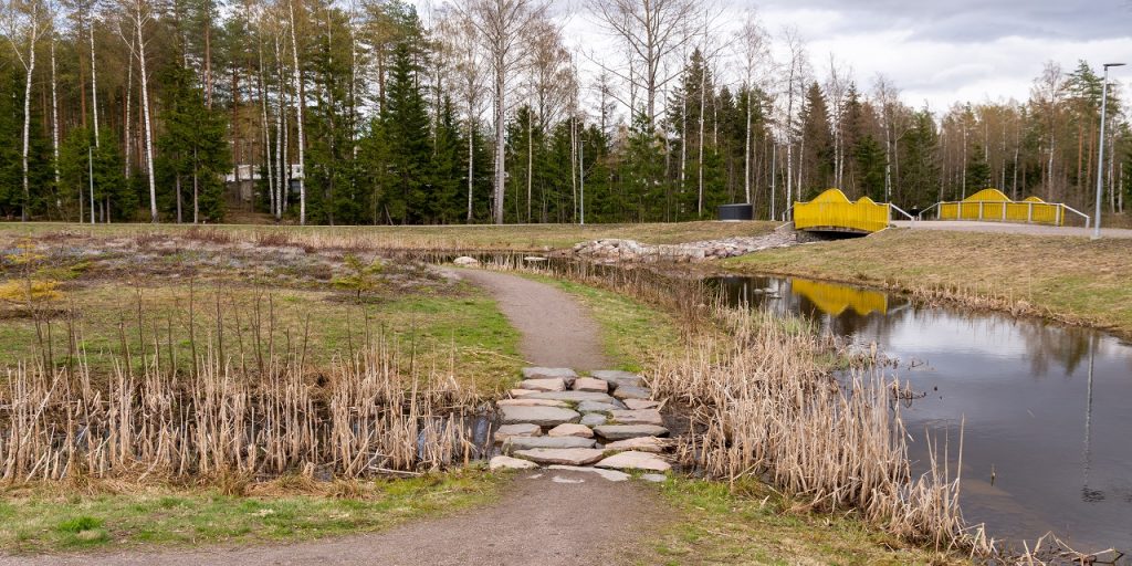 Hiekkapolku, joka kulkee eteenpäin vaihtuen ojan kohdalla isoiksi kiviksi, ympärillä on niittyä, vesikasvillisuutta ja vesiallas. Taustalla näkyy puita ja keltainen silta.