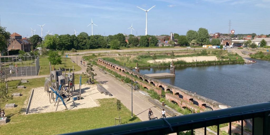 A view from a place situated high, to the park with a playground and green areas. Water area at the right, wind mills in the background.
