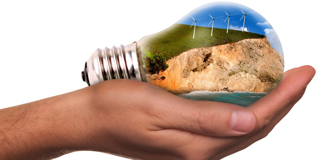 A hand holding a bulb in a palm. In the bulb, there is a photo of a cliff, with a bit of sea. On top of the cliff, there are four windmills) the sky is blue with white clouds.