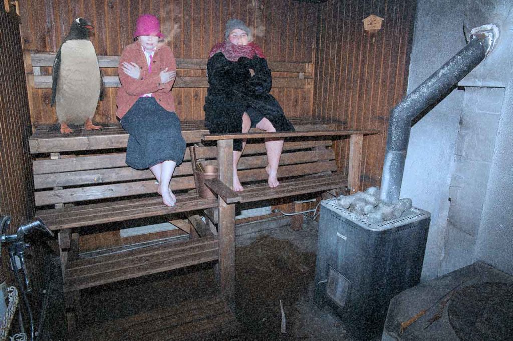 [Alt-teksti: suurehko vanhanaikainen sauna, jonka ylälauteilla istuu kaksi naista lämpimiin vaatteisiin kääriytyneinä. Heidän vieressään seisoo pingviinihahmo. Naisten hengitys höyryää.]