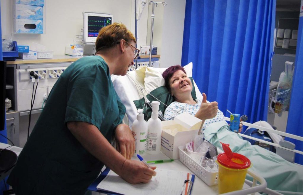 [Alt-teksti: sairaalasängyssä oleva potilas hymyilee vieressä seisovalle hoitajalle ja näyttää peukkua positiivisuuden merkkinä.]