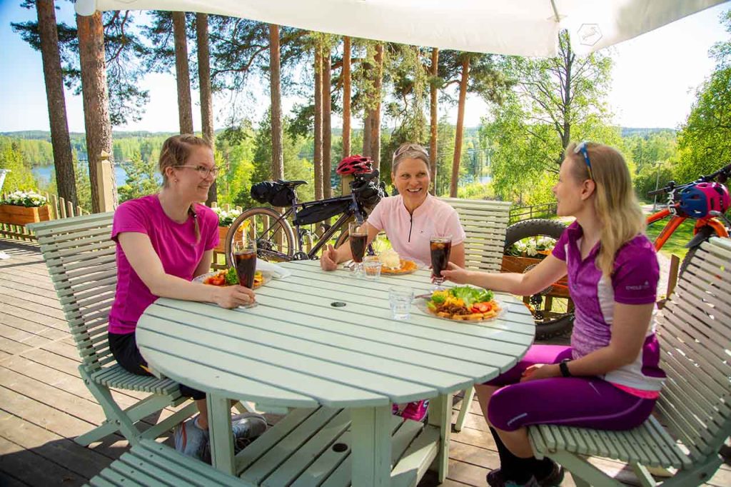 [Alt-teksti: Aurinkoinen, suuri puuterassi järvimaisemassa. Terassin pöydän ääressä istuu kolme naista, joilla on edessään ruoka-annokset ja korkeat juomalasit. Naisten pyörät ja pyöräilyvarusteet ovat terassipöydän vieressä.]