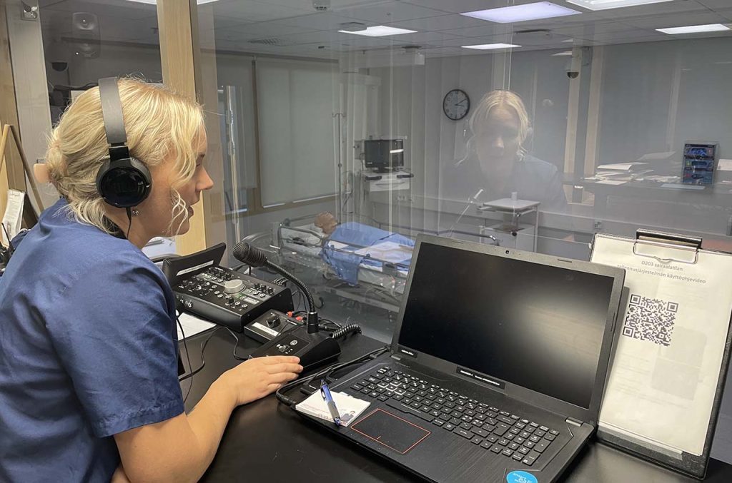 [Alt-teksti: hoitaja-asuinen nainen säätämässä tietotekniikkaa, lasiseinän takana on simulaatiotila, jossa potilassänky ja siinä makaava simulaationukke.]