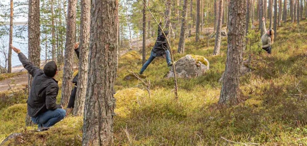 [Alt-teksti: havumetsäinen rinne, kauempana on järvenrantaa. Paksujen puunrunkojen välissä voimistelee neljä ulkoiluvaatteisiin pukeutunutta henkilöä.]