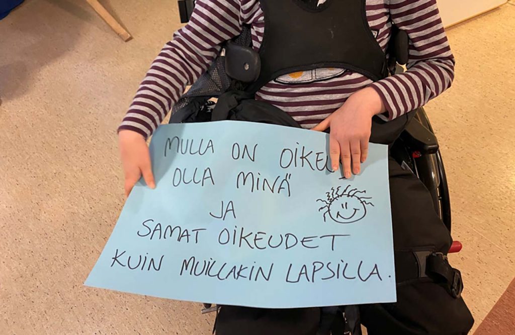 [Alt-teksti: pyörätuolissa istuva lapsi pitää sylissään suurta paperia, jossa lukee mulla on oikeus olla minä ja samat oikeudet kuin muillakin lapsilla.]