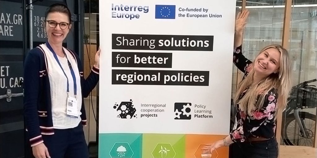 In the middle, there is a roll-up with a text saying Interreg Europe – Co-funded by the European Union. Under it, there is written Sharing solutions for better regional policies with some icons underneath. Next to the roll-up, there are two smiling women standing, each on one side of the roll-up.