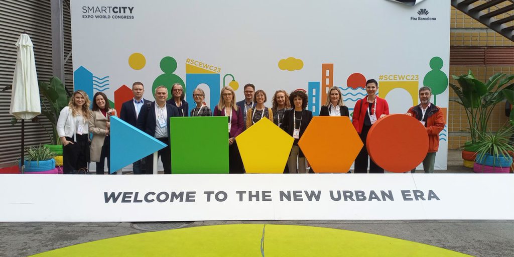 A group of standing people. In the background, there is a white board with colourful symbols related to the themes of the Smart City Expo World Congress. In the left top corner of the wall, there is a text saying Smart City Expo World Congress. In the top right corner, there is Fira Barcelona and a logo. In the front, there is a text saying Welcome to the New Urban Era, with five colourful symbols.