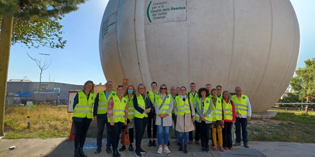 A group of people in yellow safety vests standing outdoors in front of a big whitish biogas storage tank.