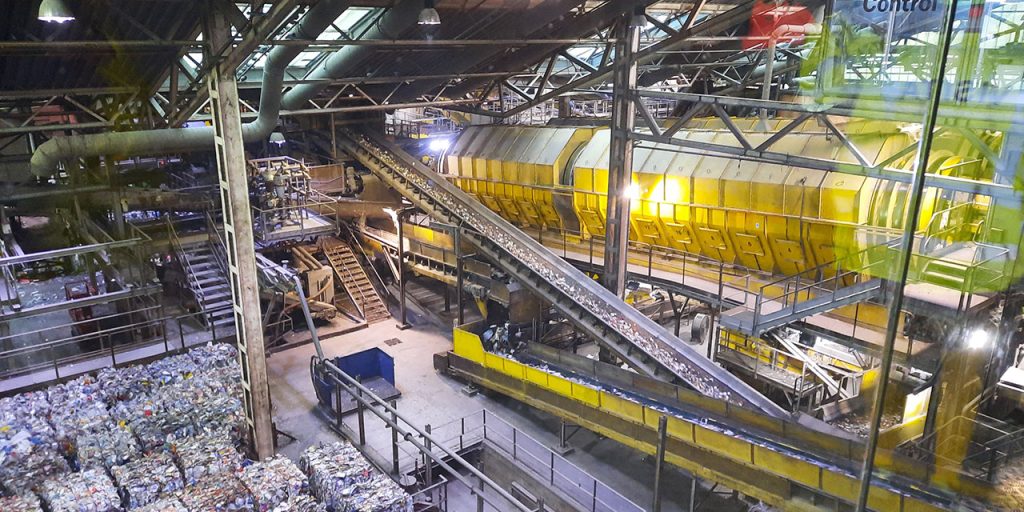 A photo from indoors of an industrial site. There are many belts and sorting machines. On the left bottom, there are packages of pressed recyclable materials.