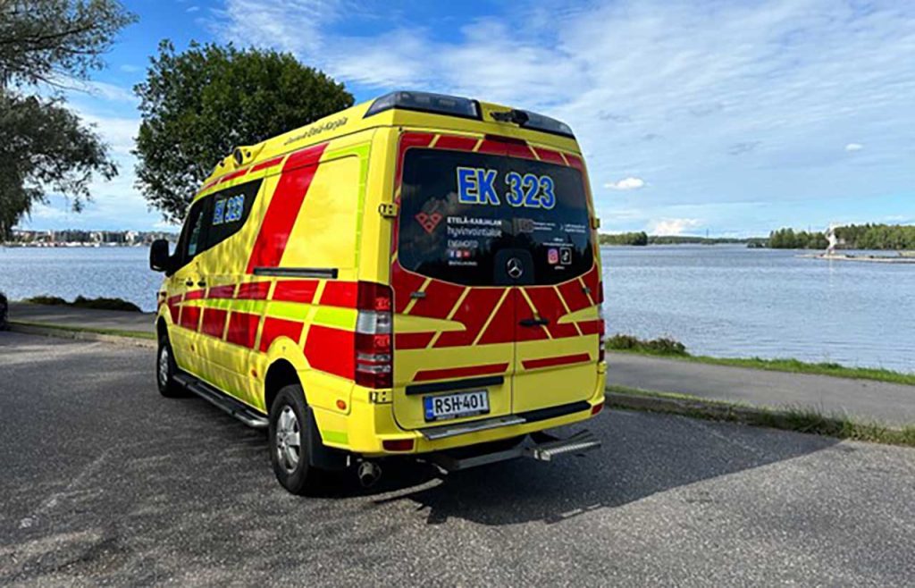 [Alt-teksti: kesäinen ranta ja parkkipaikka, jolla seisoo keltapunakuvioinen ambulanssi.]