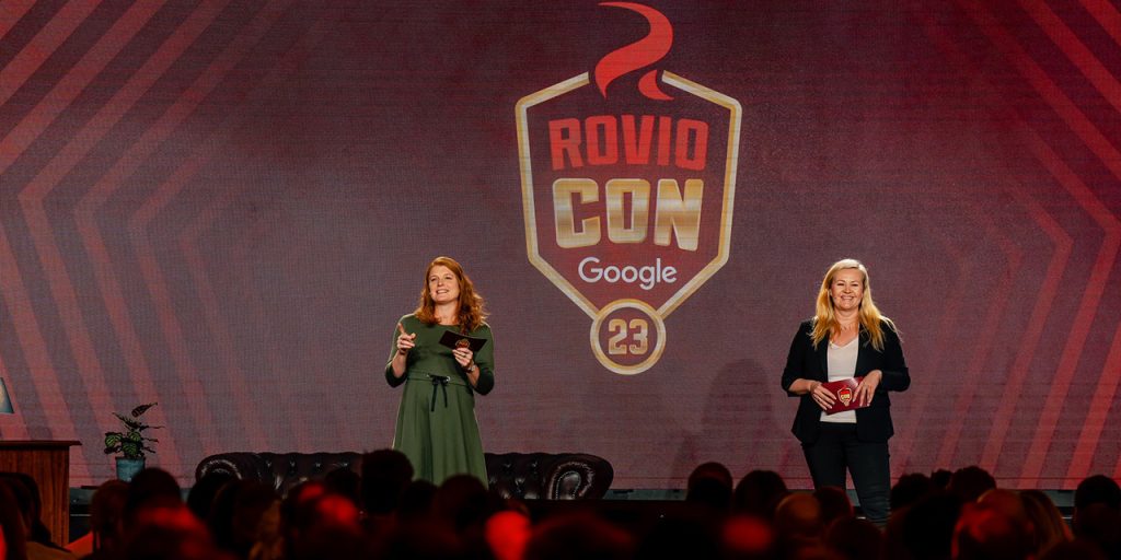 Two female figures standing in front of big, red and gold coloured logo saying RovioCon Google 2023.