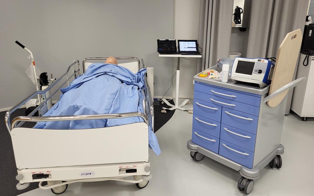 [Alt-teksti: simulaatiotilassa on sairaalasänky, jossa potilasnukke, sekä erilaisia hoitotarvikkeita sängyn vieressä työtasolla, monitoreita ynnä muuta.]