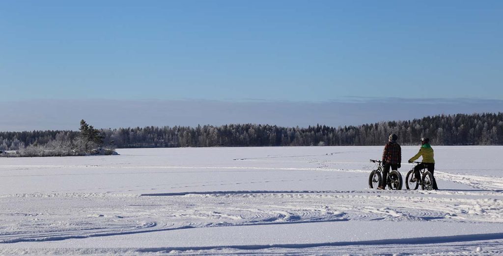 [Alt-teksti: jäätynyt järvenselkä lumen peitossa, lumikentällä kaksi henkilöä maastopyörien kanssa.]