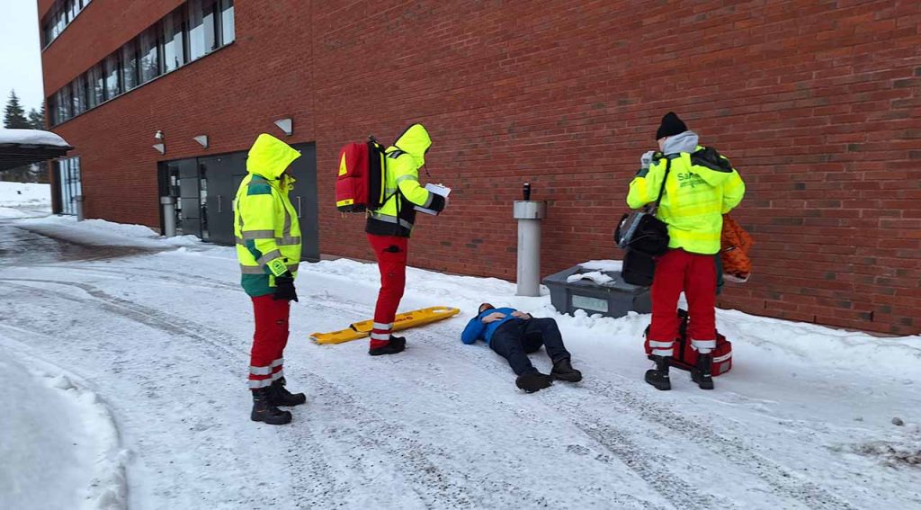 [Alt-teksti: ryhmä ihmisiä rakennuksen seinän vieressä ulkona. Maassa lumella makaa yksi henkilö, muut ovat saapuneet häntä auttamaan.]