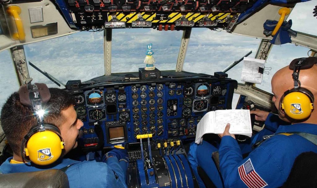 [Alt-teksti: lentokoneen ohjaamo, jossa istuu kaksi miestä kuulokkeet korvilla, ikkunoista avautuu näkymä pilvien yläpuolella.]