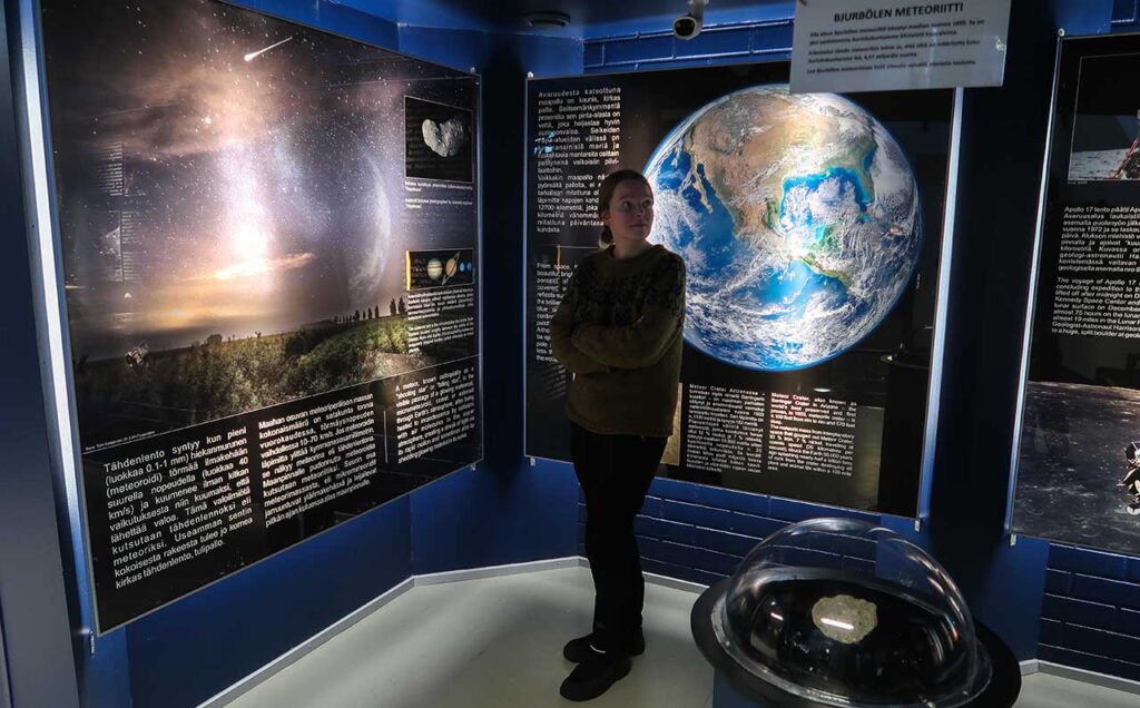 [Alt-teksti: museon näyttelyseinä, jossa suuria kuvia maapallosta, tähtitaivaasta ynnä muuta sekä paljon infotekstejä, näyttelyseinän edessä seisoo nuori nainen.]