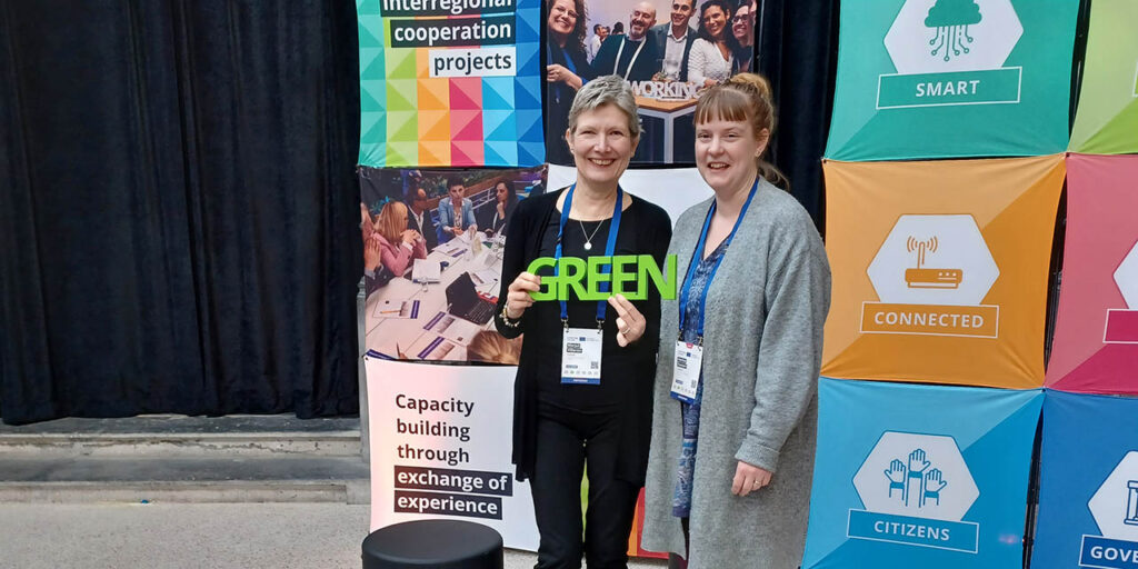 Two people standing in the front of a colourful background. The other one is holding a sign with the word GREEN.