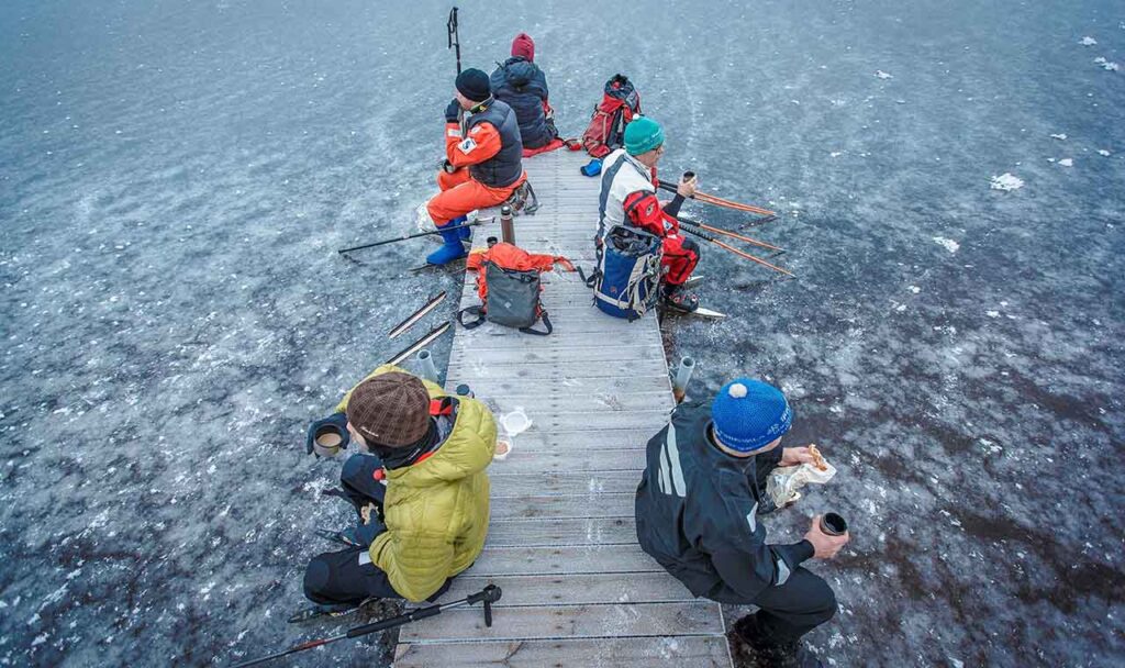 [Alt-teksti: jäätyneen järven rannalla pitkä laituri, jolla istuu viisi retkeilijää nauttimassa eväitään.]