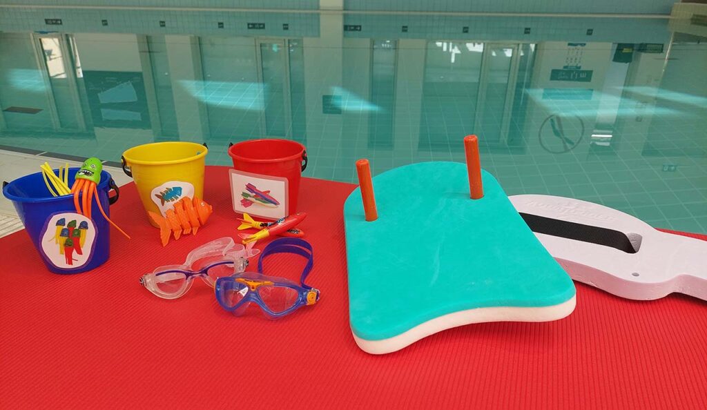 [Alt-teksti: uima-altaan reunalla punaisen maton päällä sininen, keltainen ja punainen hiekkaämpäri, joissa on erilaisia sukellusleluja. Kahdet uimalasit, uimalauta ja vesijuoksuvyö.]