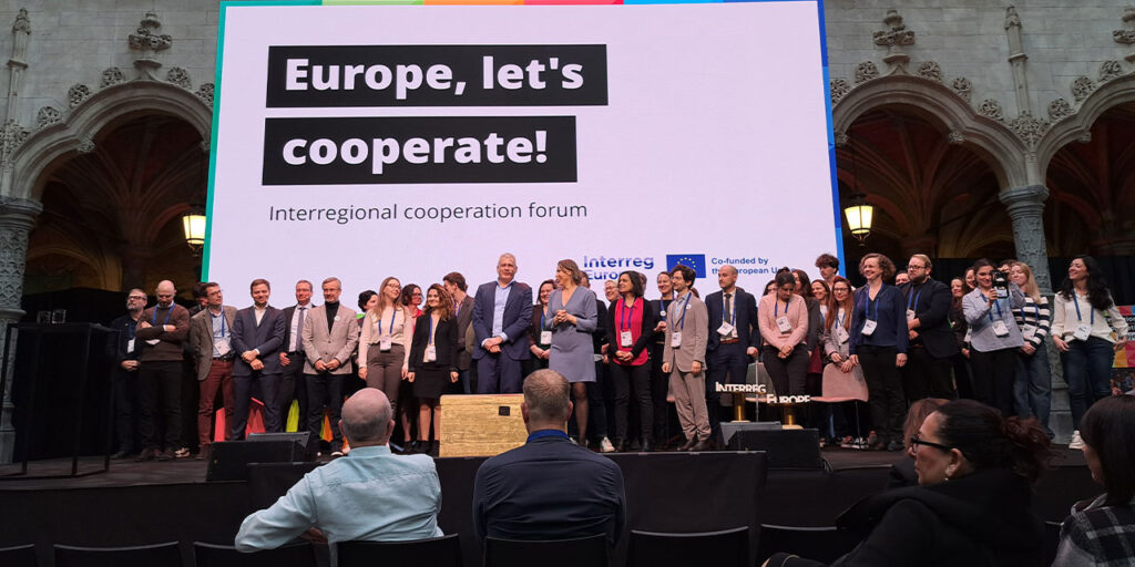 A photo indoor, a group of people standing on a stage. Behind them is a big screen showing the text of Europe, let’s cooperate! – Interregional cooperation forum.
