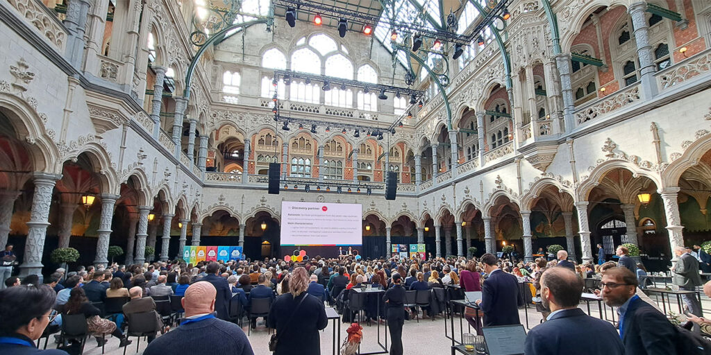 A photo indoor, an old beautifully decorated building, with hundreds of people sitting and facing the big screen placed at the far back center of the room.