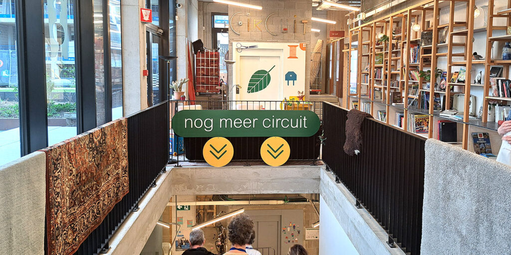 A photo indoor with a sign “nog meer circuit”, and the name of CIRCUIT on the wall at the back, made of pieces of various products and tools. On the right, there are shelves with books. On the left, there are windows. In the middle is a partly visible staircase downstairs.
