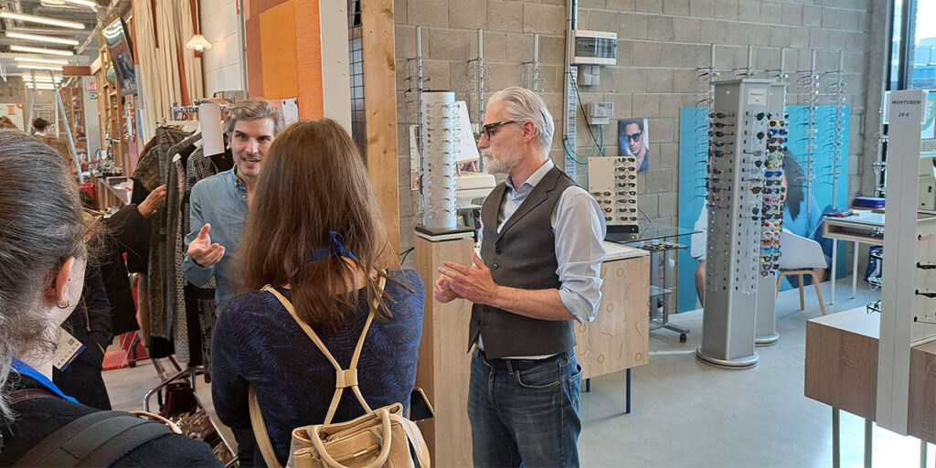 A photo indoor of a group of people listening to a man who explains something. On the right, there is a shop with glasses and sunglasses displayed on several stands.