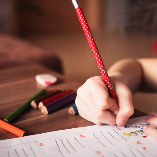[alt-teksti: Lapsi piirtää paperille pöydän ääressä, jolla on eri värisiä puuvärikyniä.]