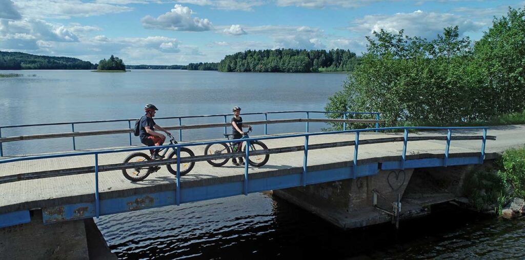 [Alt-teksti: kesäisenä päivänä suuri järvenselkä ja lähempänä veden ylittävä silta, jossa on levähtämässä kaksi pyöräilijää.]