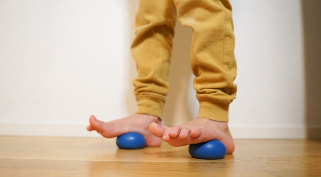 [Alt-teksti: lapsen jalat, kummankin jalkaterän alla on litistyvä sininen pieni pehmeännäköinen pallo.]