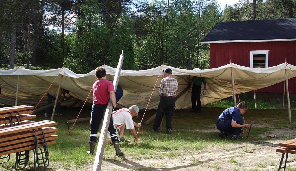 [Alt-teksti: kesäinen perinnepiha, punamultaseinäinen pieni rakennus taustalla, kuuden ihmisen ryhmä pystyttää telttakatosta.]