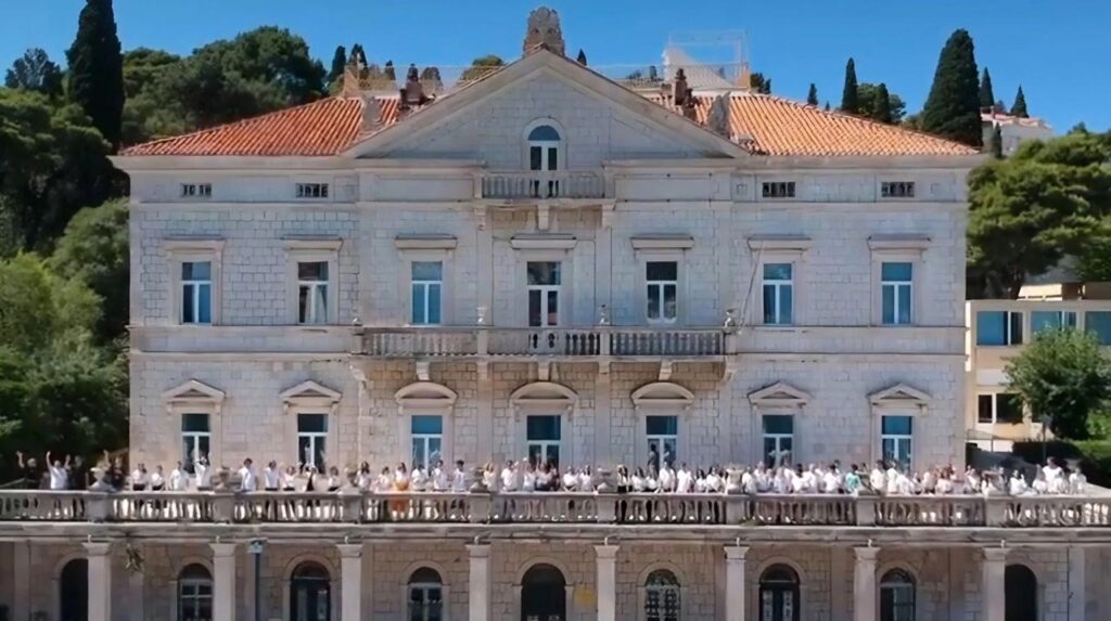 [Alt text: a very big and old-looking building, in front of it a terrace with many people waving hands.]