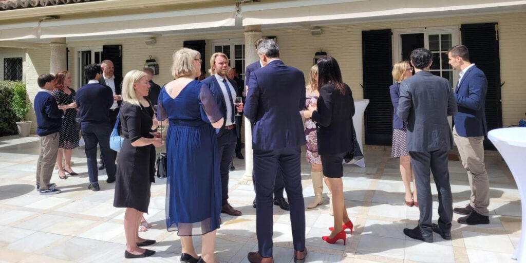 [Alt text: a group of smartly dressed people talking in front of a mansion.]