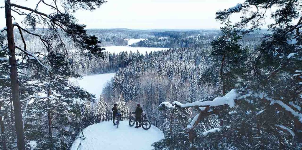 [Alt-teksti: talvinen maisema, paljon lumisia puita, puiden keskellä on näköalatasanne, johon on pysähtynyt kaksi talvipyöräilijää.]