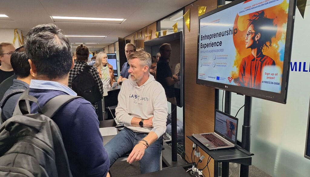 [Alt text: people gathered around in the hall to discuss and meet each other, you can also see posters, computers etc.]